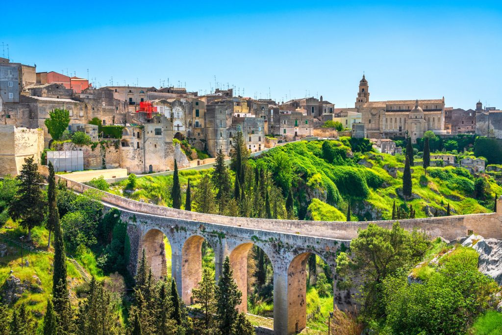 Panoramic shot of Gravina in Puglia featuring the massive two-tier stone bridge. In the background, the ancient white stone buildings of the town sit on the edge of a steep limestone cliff.