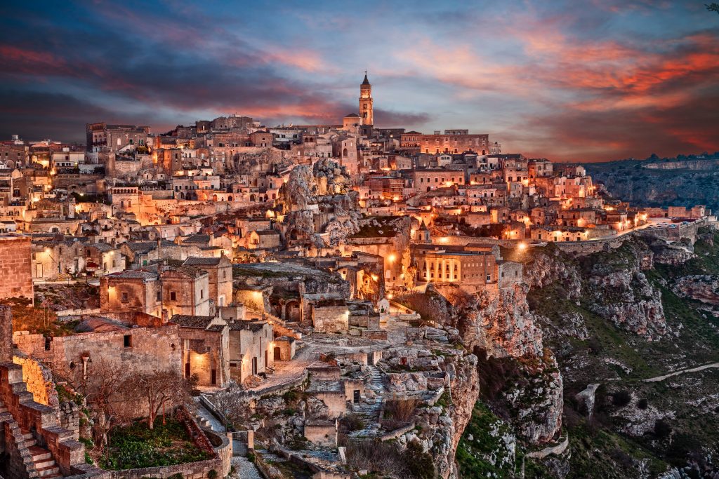 A wide panoramic shot of the Sassi di Matera, showing the ancient cave dwellings and stone houses built into the limestone cliffs. The city rises in layers with narrow streets and cave openings under a clear sky