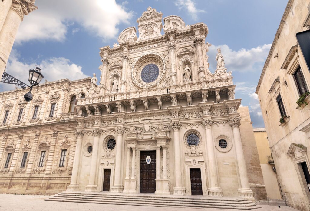 The ornate facade of the Basilica of Santa Croce in Lecce, Italy. The golden limestone is intricately carved with sculptures of angels, animals, and floral patterns, featuring a grand central rose window.