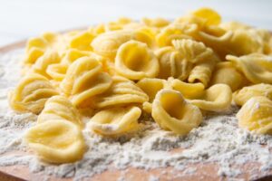 A close-up of fresh, handmade orecchiette pasta resting on a wooden surface dusted with semolina flour.