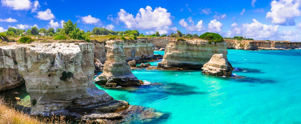 High-angle shot of the 'Faraglioni di Sant'Andrea' near Melendugno. The bright white rock formations contrast sharply with the vibrant emerald and deep blue waters under a bright Italian sun.